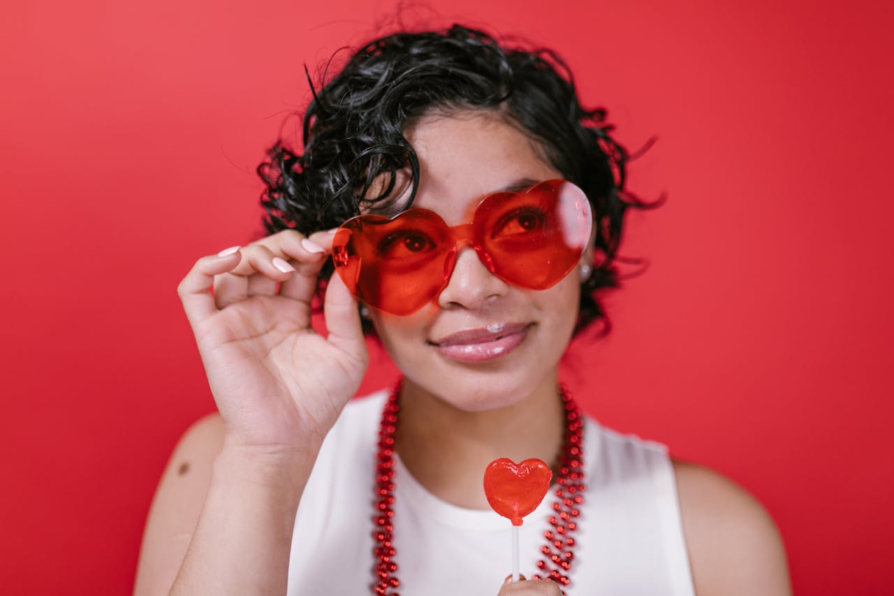 gallery-img-05 Woman in red sunglasses and necklace smiles while holding a heart-shaped lollipop against a red background.