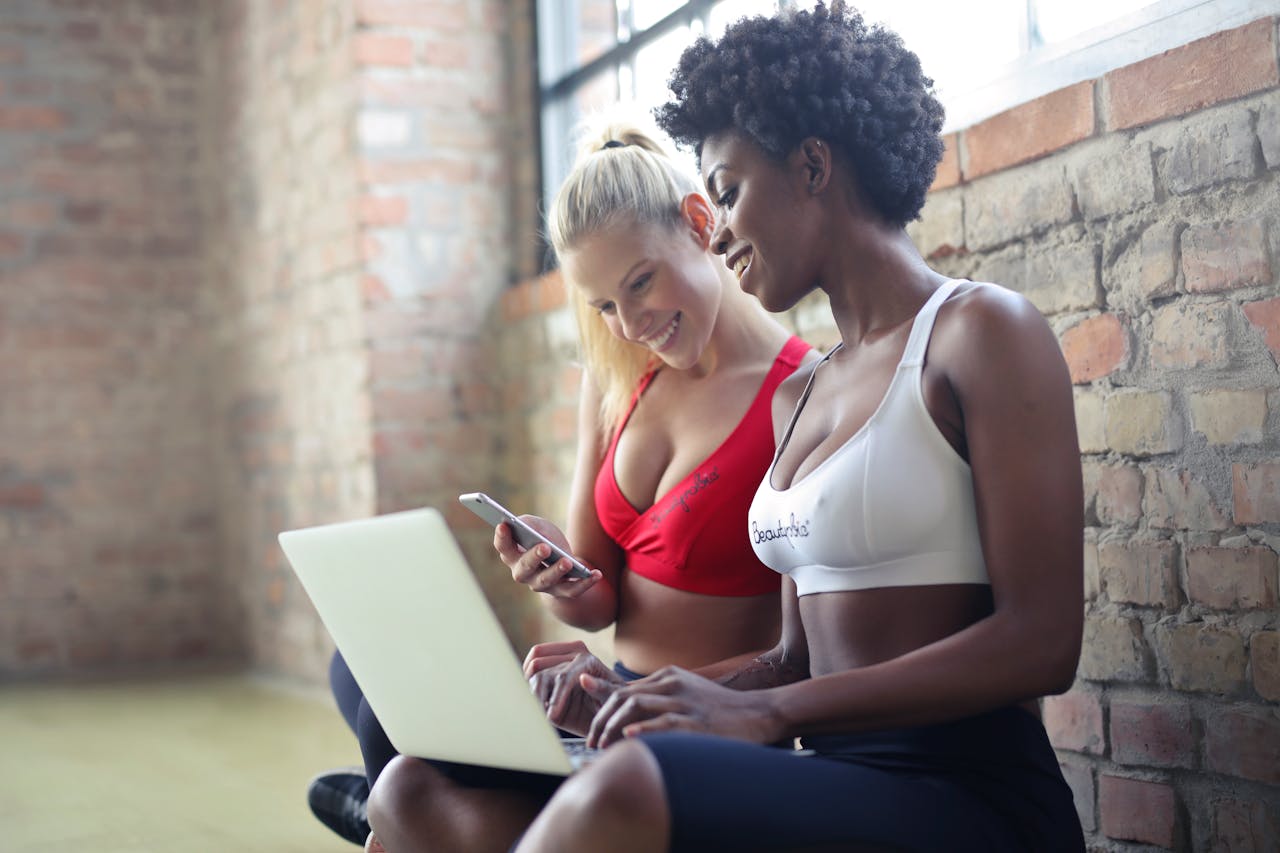 extra-programs-img Two women in sportswear enjoy leisure time, sharing a laptop and smartphone in a gym setting against a brick wall.